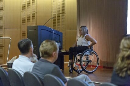 alycia delivering a speech on a stage in her wheelchair sitting next to a podium