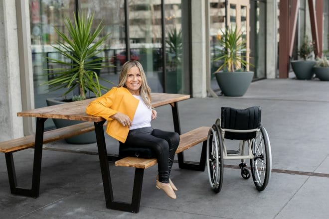 alycia sitting on a table bench next to her wheelchair in a yellow jacket