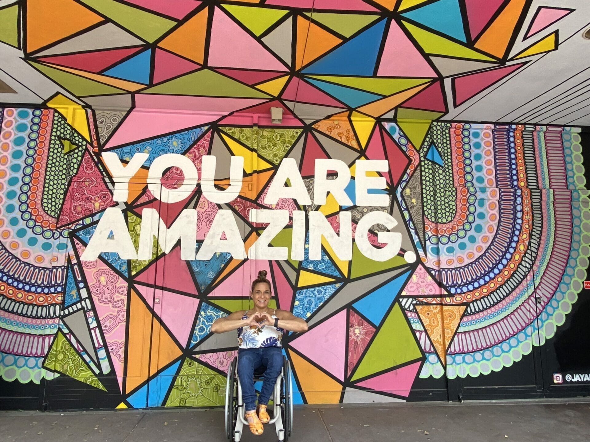 alycia sitting in fron of a mural in phoenix arizona that says you are amazing while she is making the heart symbol with her hands