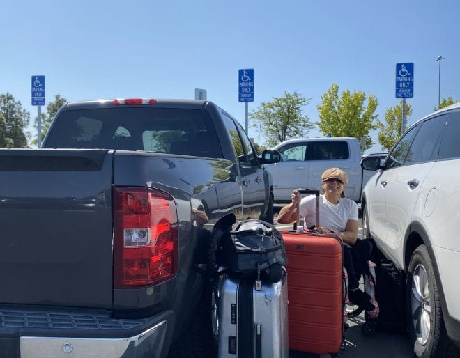 alycia with suitcases and luggage between a truck and an suv sitting in her wheelchair in a handicap parking spot 
