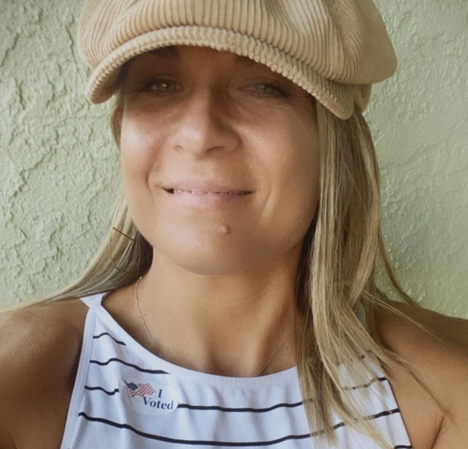 ID: Alycia wearing a brown hate and white striped shirt with her I voted sticker on her shirt smiling.