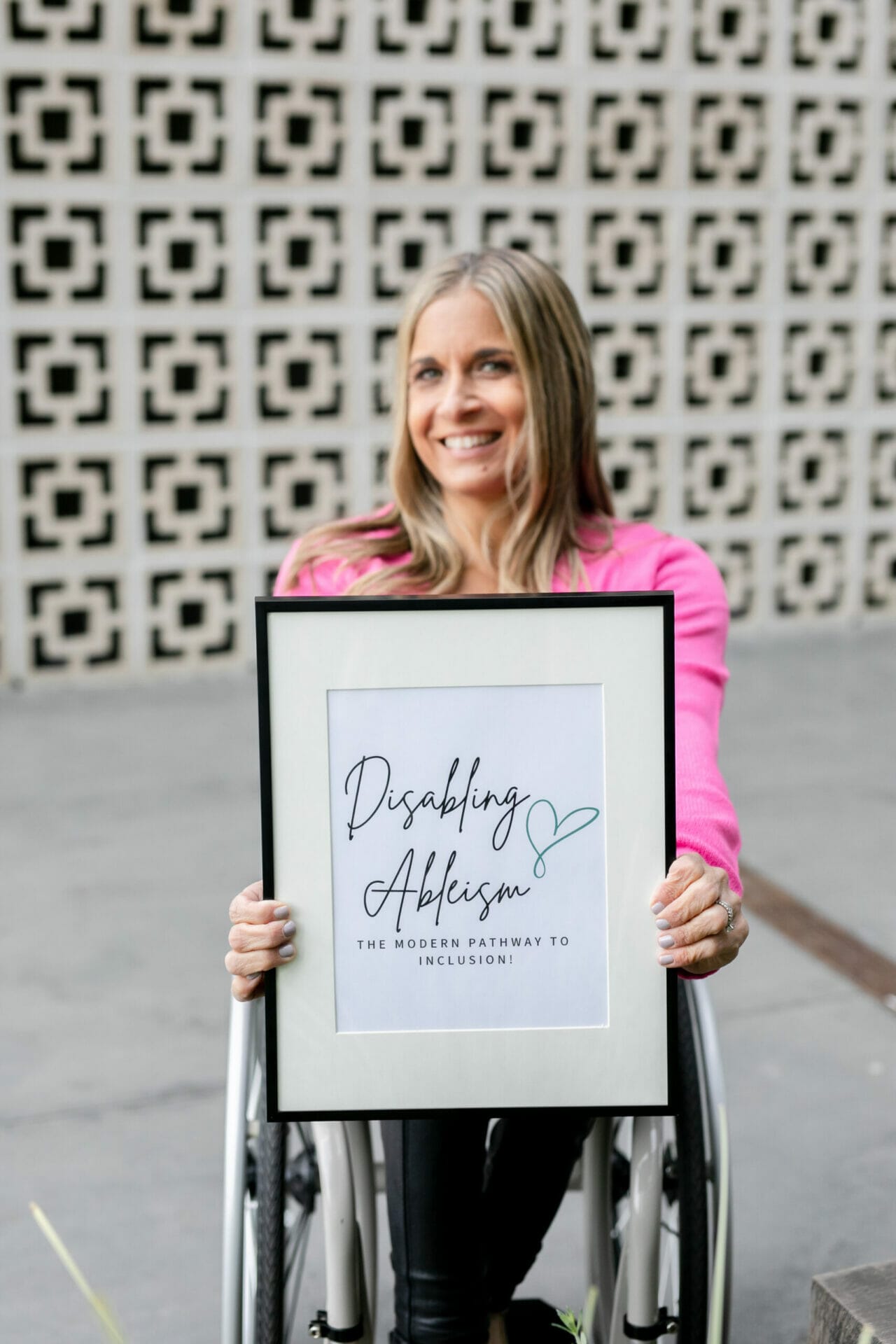 Alycia wearing pink sitting in wheelchair holding black picture frame that says disabling ableism the modern pathway to inclusion.