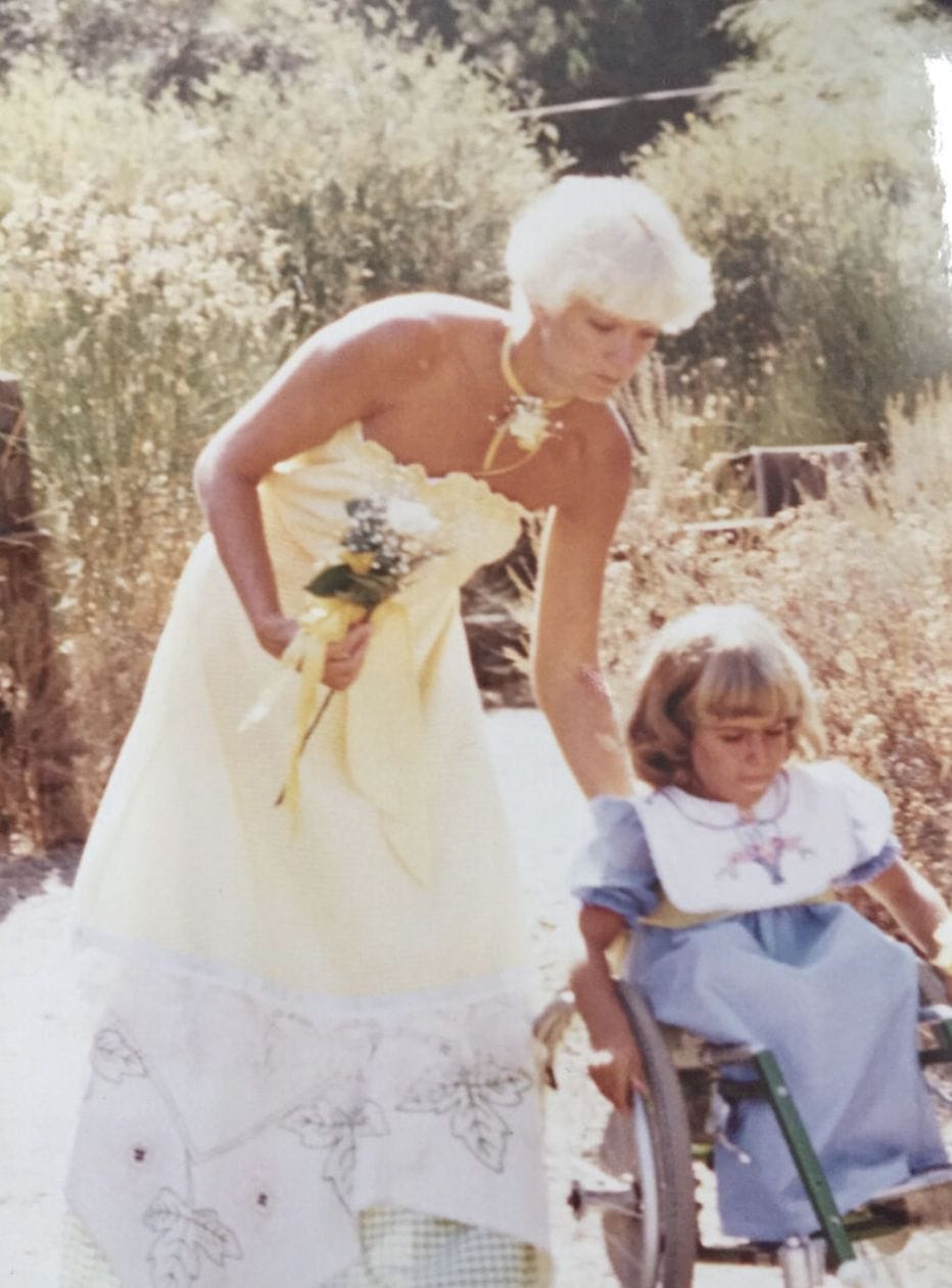 ID: Alycia and her mom in a wedding pushing her wheelchair down a rocky road carrying flowers