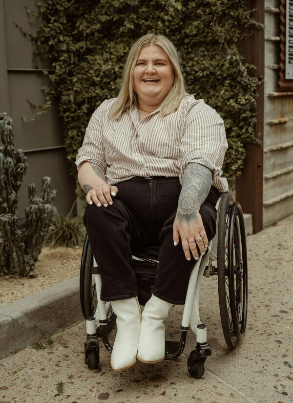 maegen sitting in her wheelchair smiling wearing a striped shirt blue jeans and white boots in front of an ivy bush