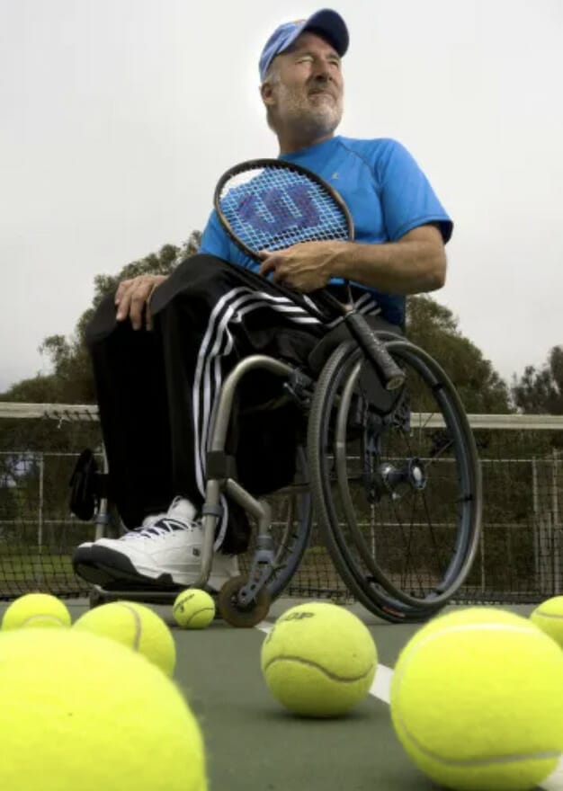 brad parks sitting in his wheelchair on a tennis court holding a tennis racquet with tennis balls on the ground in front of him