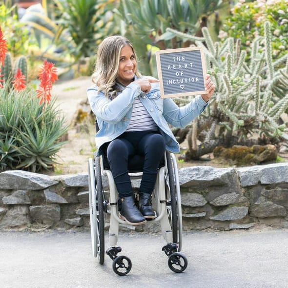 alycia sitting in her wheelchair in front of a succulent garden pointing at a frame with words in it that say the heart of inclusion