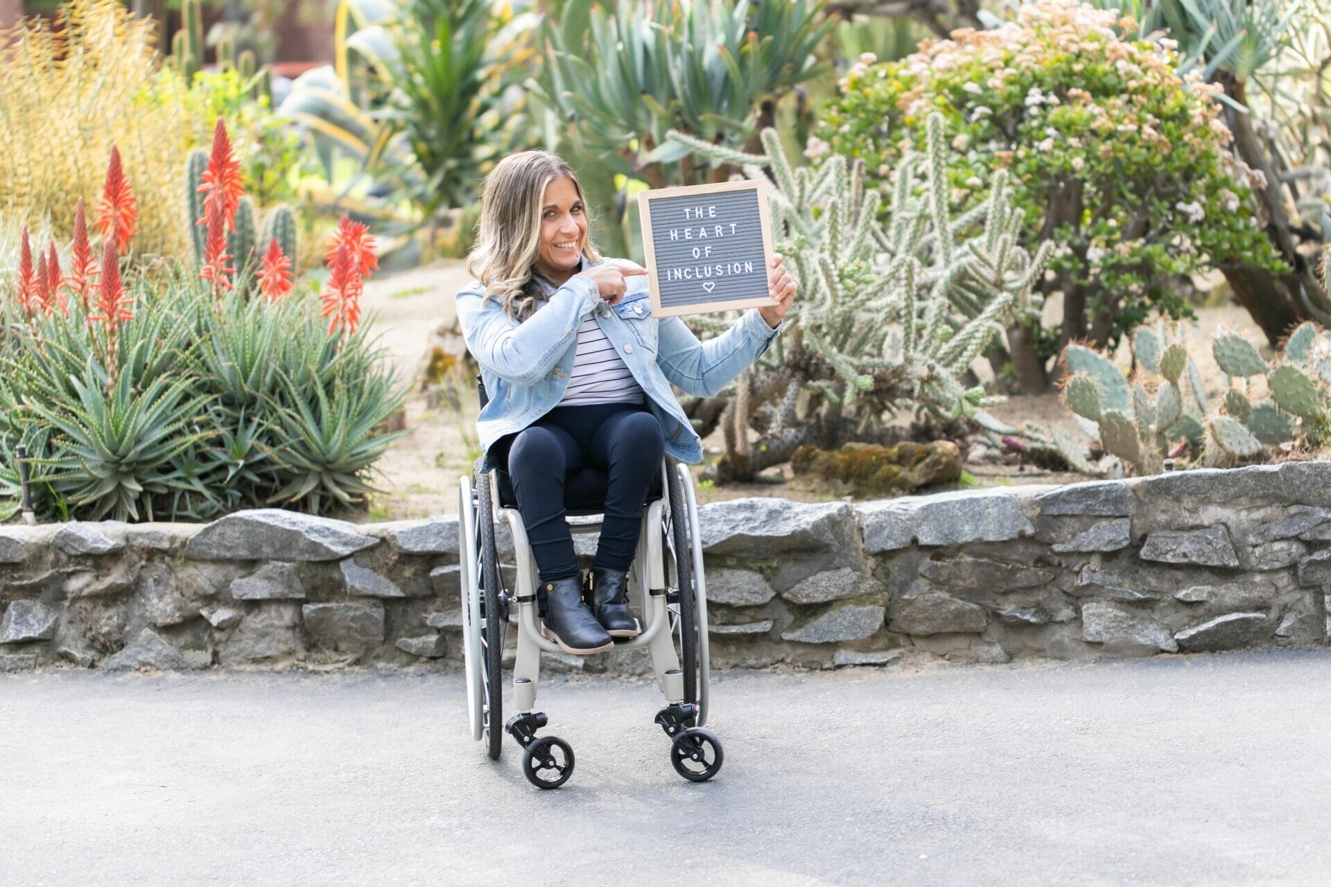 Alycia sitting in her wheelchair in front of green cactus, wearing a jean jacket and black pants and boots holding a sign that says The Heart of Inclusion.