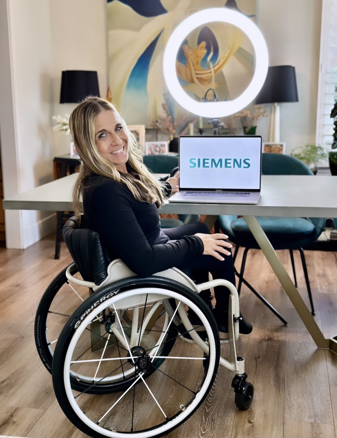 ID: Alycia wearing all black sitting in her wheelchair at her home studio in front of a table with a laptop showing the Siemens logo.