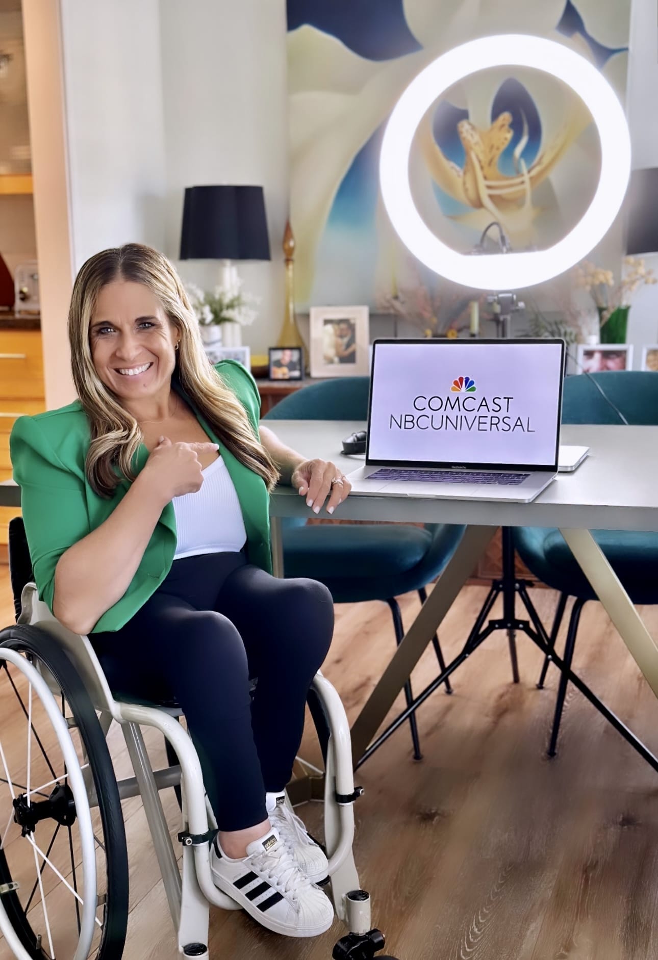 Alycia sitting in her studio in front of her computer after her COMCAST/NBC keynote. She is wearing a green jacket and black pants and tennis shoes pointing at her computer with the COMCAST/NBC logo and ring light behind her.