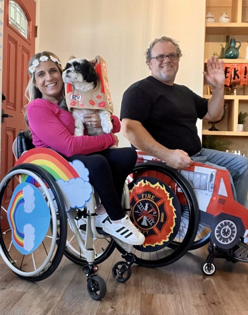 Alycia sitting in wheelchair wearing rainbow cloud costume and Daisy flower crown and Marty sitting in wheelchair wearing firefighter costume from Targets adaptive costume line pointing at their red logo.