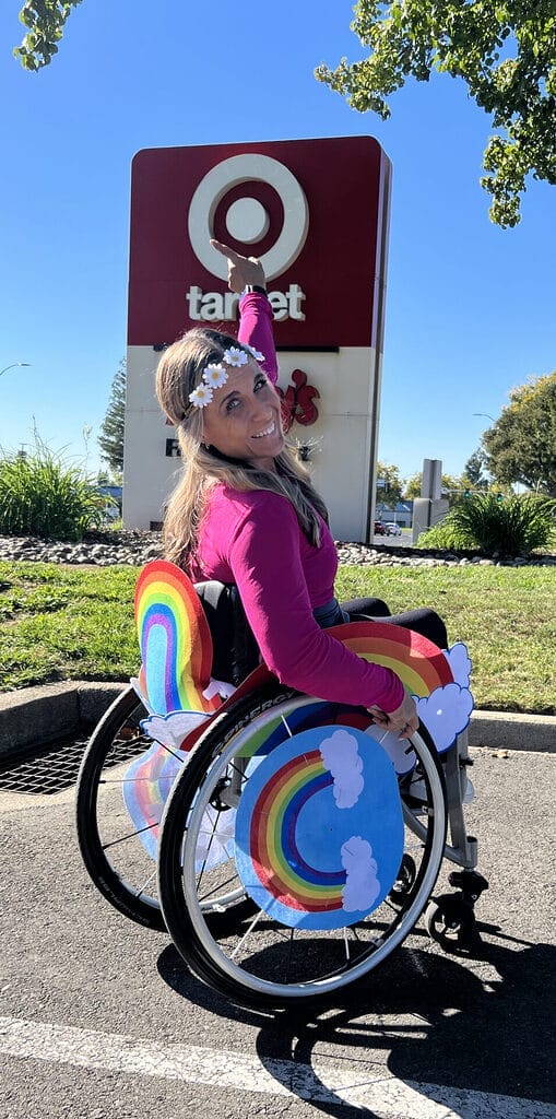 Alycia sitting in wheelchair wearing rainbow cloud costume and Daisy flower crown from Targets adaptive costume line pointing at their red logo.