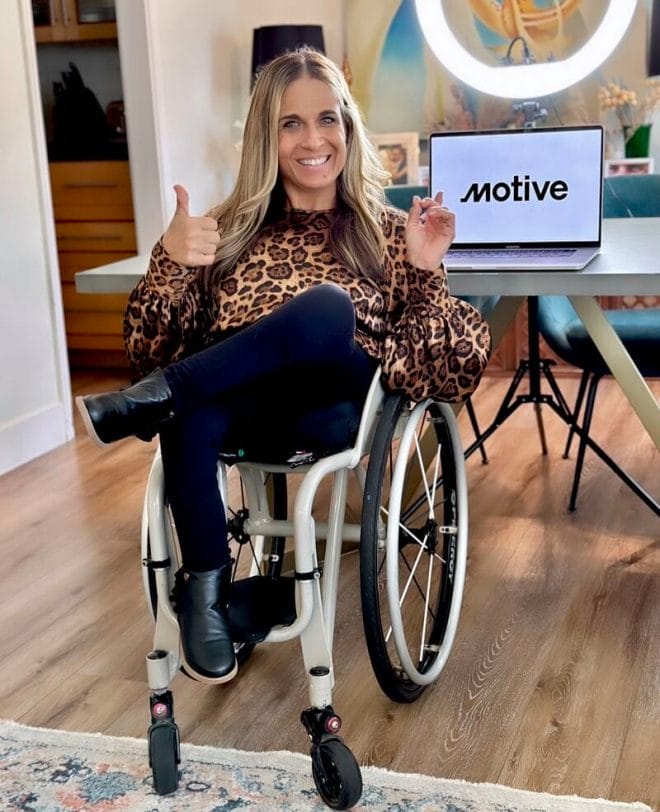 alycia sitting in her wheelchair wearing a leopard skin blouse giving a thumbs up and pointing at her laptop with the motive logo showing on it