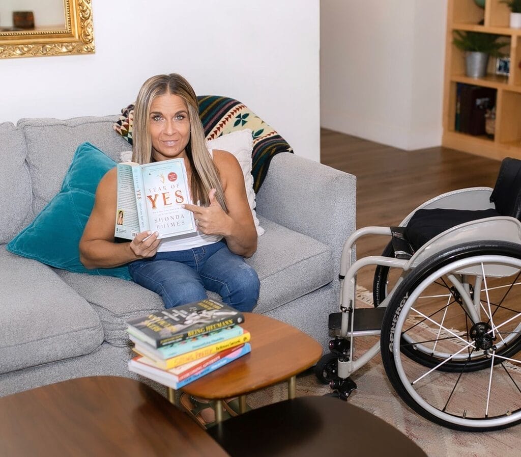 alycia sitting on a couch with her wheelchair next to her holding the book yes by shonda rhimes