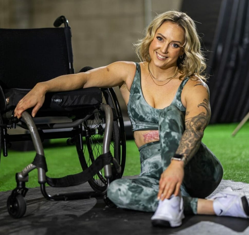 nikki walsh sitting on the ground next to her wheelchair resting her arm on the wheelchair seat