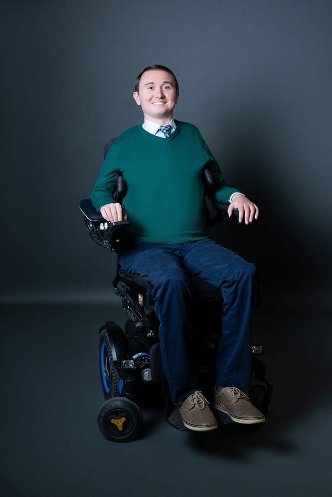 josh basile sitting in his power wheelchair wearing a green sweater and blue pants