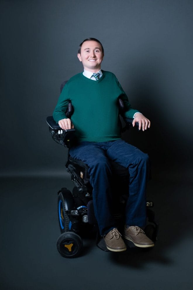 josh basile sitting in his power wheelchair wearing a green sweater and blue pants