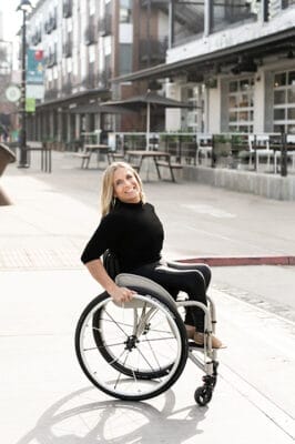 alycia anderson sitting in her wheelchair wearing a black top and pants on a city sidewalk