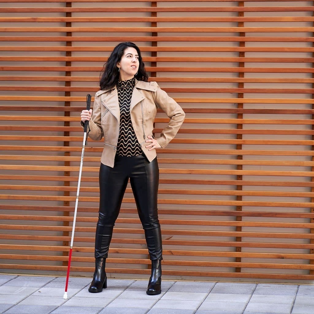 catarina rivera standing in front of a slat wall holding her cane wearing a tan leather jacket and black tights