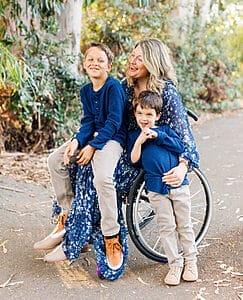 lindsay ulrey sitting in her wheelchair smiling wearing a blue patterned dress with her sons one sitting on her lap and one standing and leaning against her