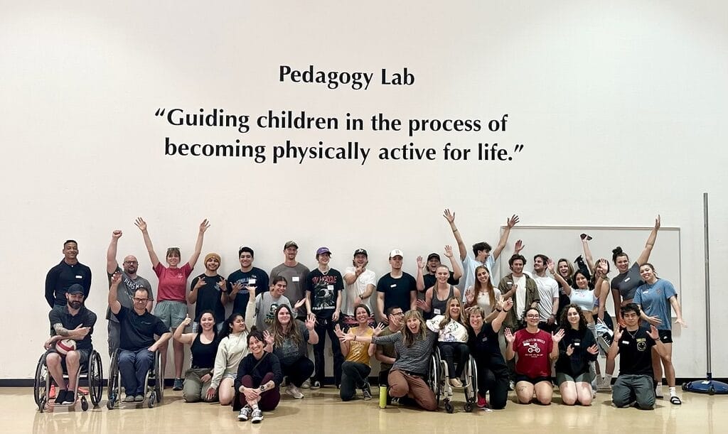 alycia posing with a large number of students in the yolo hall sports gym with a saying on the wall pedagogy lab guiding children in the process of becoming physically active for life