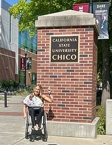 alycia sitting in her wheelchair pointing up at a pillar that has a plaque that says california state university chico