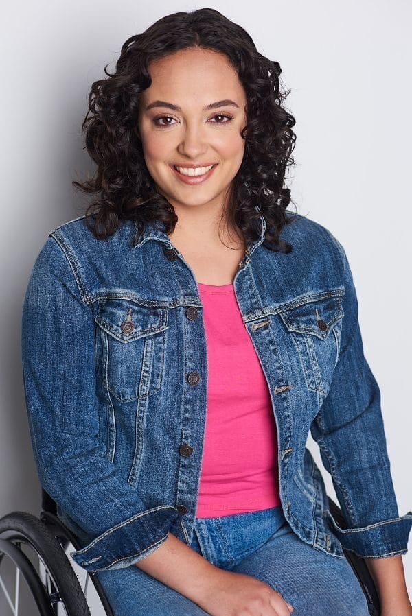 joci scott smiling with long curly dark hair sitting in her wheelchair wearing a blue jean jacket and pants with a pink shirt