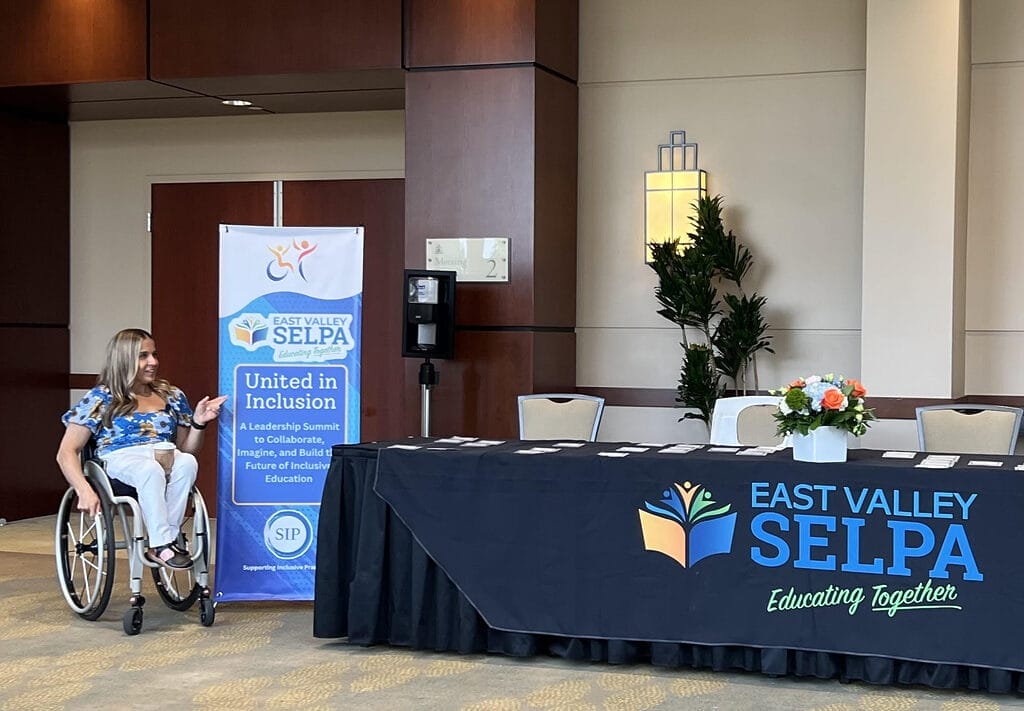 alycia sitting at the welcome desk for the east valley selpa pointing at some marketing that says united in inclusion