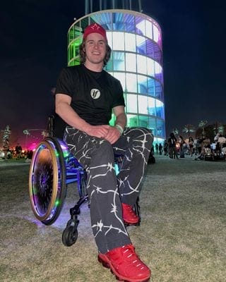 mason branstrator sitting in his wheelchair in front of a glowing building of some sort with lights on his wheels and lightning bolt pants