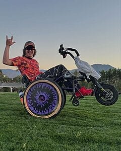 mason branstrator popping a wheelie in his wheelchair on a grassy lawn with a motorized wheel attachment giving a i love you hand sign