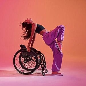 kaylee bays performing a dance move where she is standing in front of her wheelchair reaching backwards and holding the wheels behind her leaning back fully with one leg resting on the other legs knee with an image background gradient of orange to pink kaylee is wearing a black top and baggy purple pants