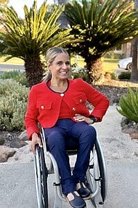 alycia sitting in her wheelchair wearing a red blazer in front of seco palms with one hand on her wheel and the other on her hip