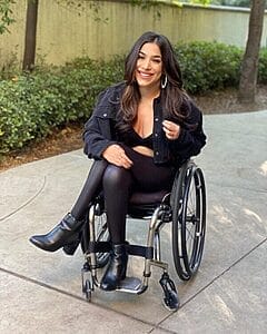 steph aiello sitting in her wheelchair wearing an all black outfit with jacket leggings and boots she has her legs crossed and her hand on one knee she is smiling