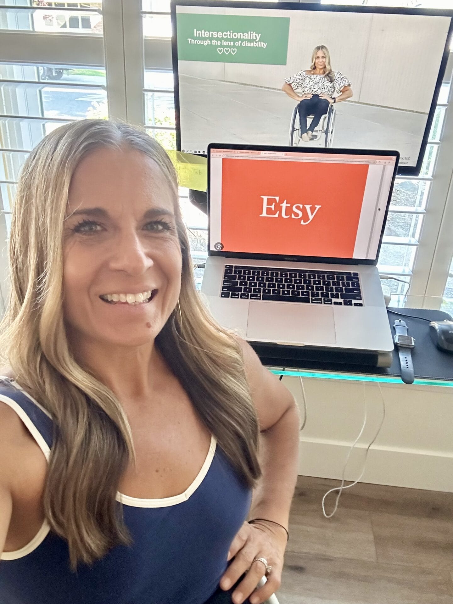 alycia sitting in front of her virtual studio with etsy logo on one monitor and her interstionality through the lens of disability presentation on another smiling doing a selfie with her hand on her hip