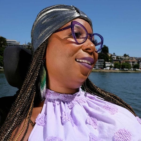 andraéa lavant wearing a lavender blouse with big glasses smiling from a side profile headshot beside a lake