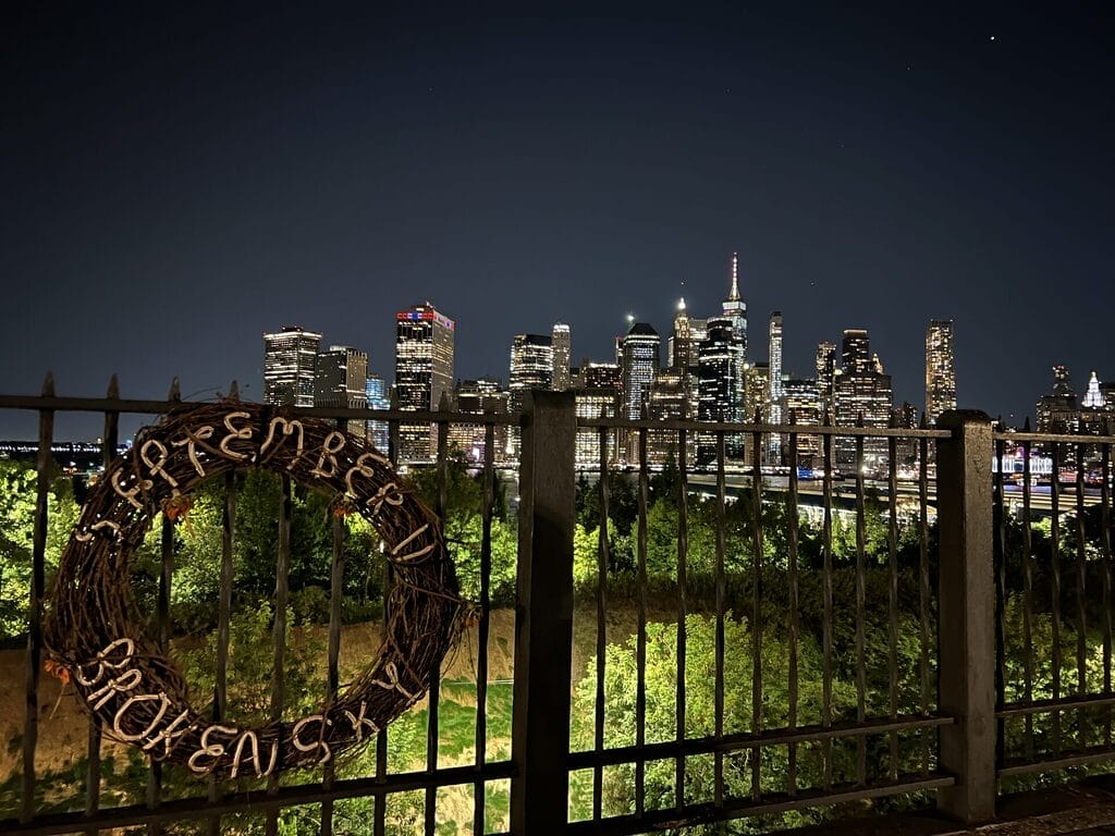 a picture of the new york sky line from the promenade in brooklyn looking at the new world trade with a wreath on the fence in the foreground that says september eleven broken sky
