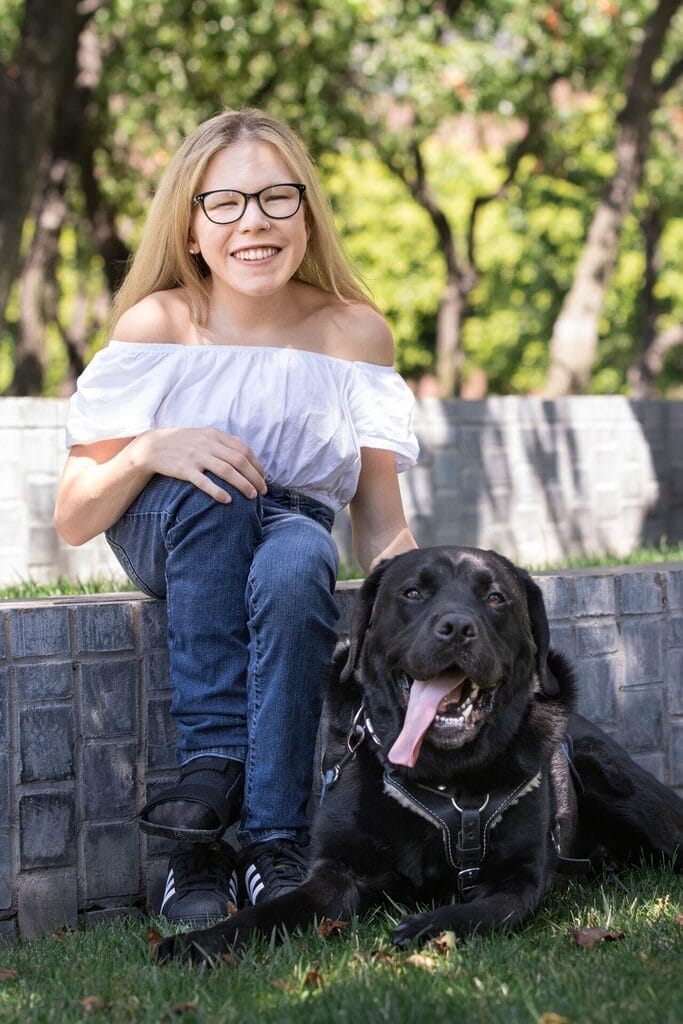 cassidy huff sitting on a wall looking foward smiling while petting her dog beside her who is also looking forward laying next to her with his tongue hanging out