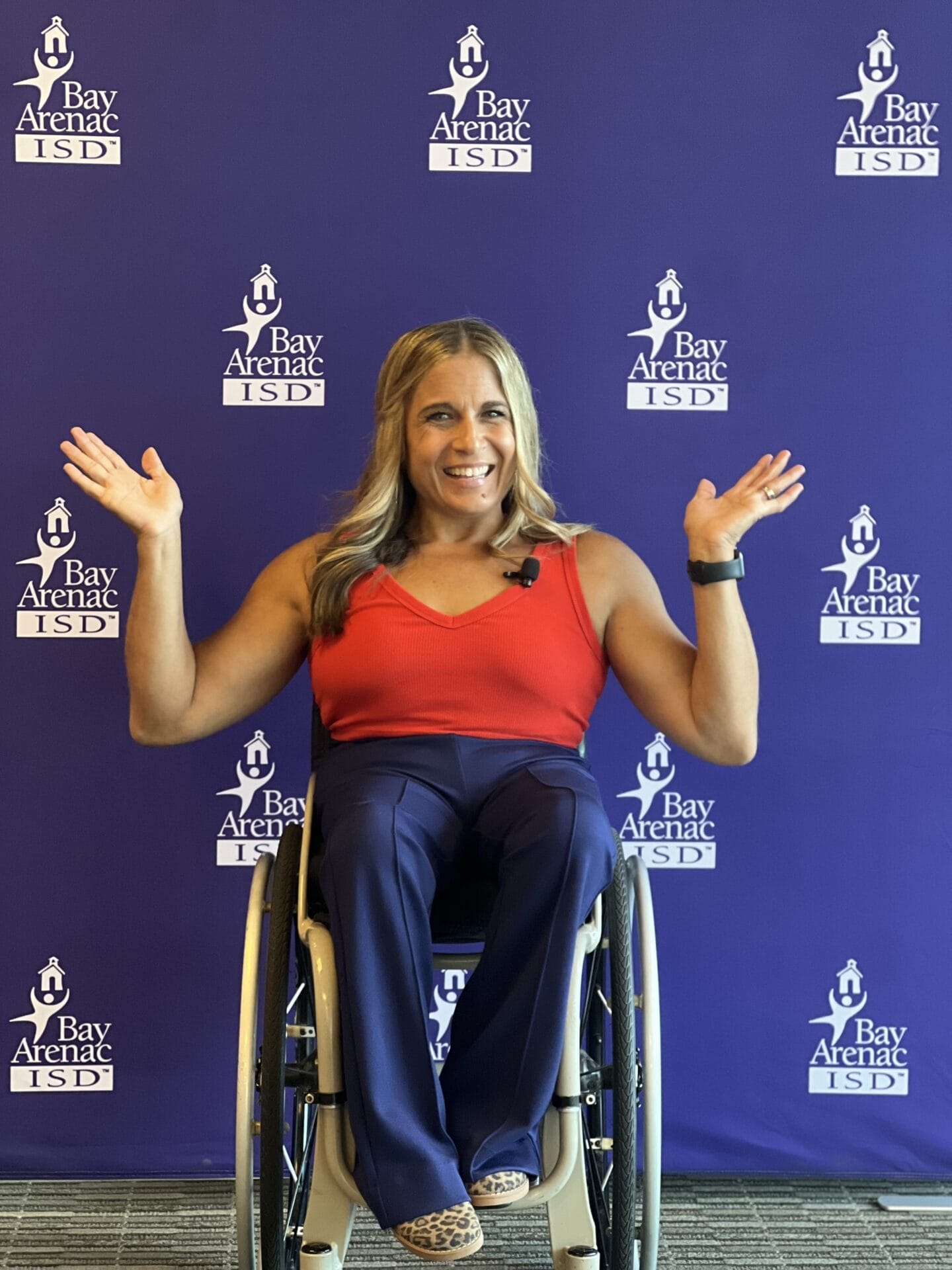 alycia sitting in her wheelchair in front of the bay arenac isd logo wall holding her hands up and open smiling