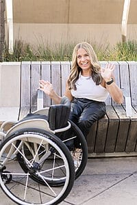 alycia sitting on a bench next to her wheelchair waving her hands and smiling