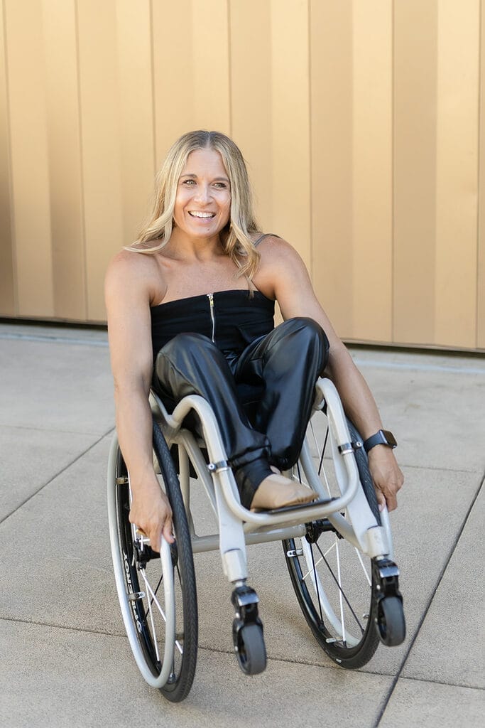 Alycia wearing all black doing a pop a wheelie in wheelchair near a gold wall