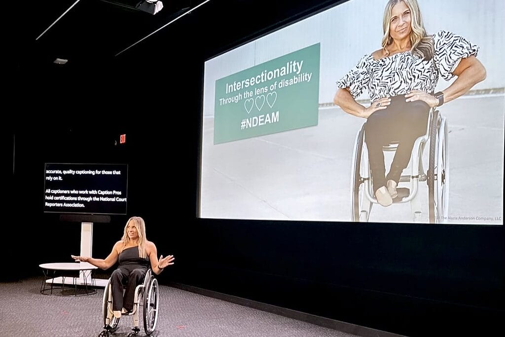 alycia on stage at the bronson theater netflix stage with her hands spread out and open under a screen with her on it that reads intersetionality through the lens of disability hashtag n d e a m