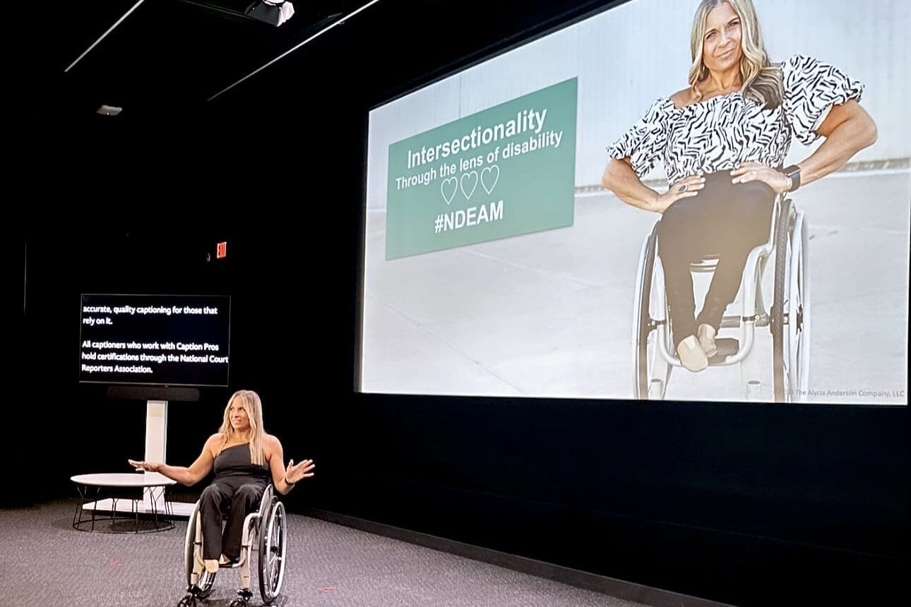 alycia on stage at the bronson theater netflix stage with her hands spread out and open under a screen with her on it that reads intersetionality through the lens of disability hashtag n d e a m