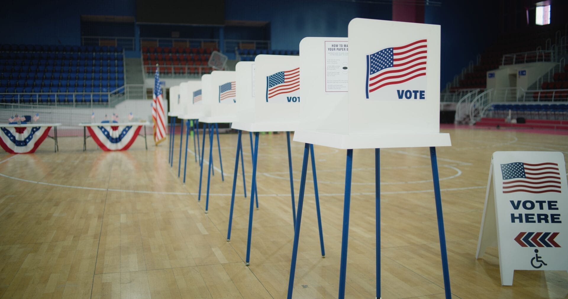 gymnasium set up with voting booths and voting monitor tables including a sign with american flag stating vote here with arrows pointing and disabled symbol
