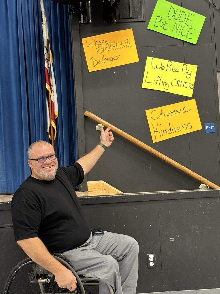Marty wearing black sitting in wheelchair near signs that encourage inclusion