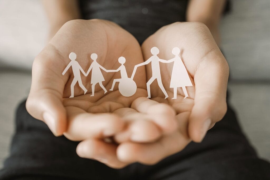 wooden cutouts of men and women on both sides of a person with a disability in a wheelchair all connected laying in the palms of open hands