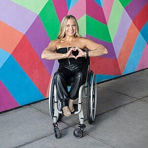alycia sitting in her wheelchair in front of a colorful star mural making the sign of a heart with her hands