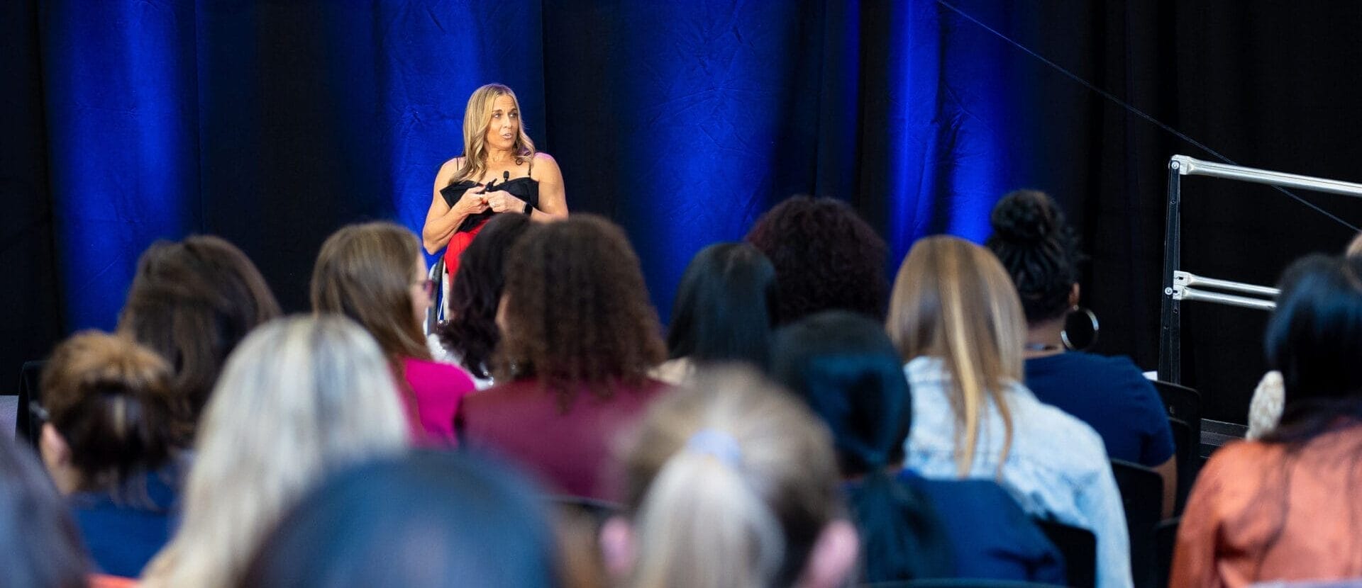 alycia on stage under the women empowerment event logo with sponsors to her left is a ramp on to the stage and a number of women are in the crowd in front of her