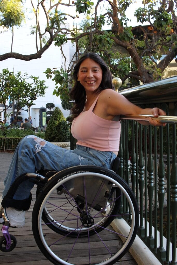 bella adlah sitting in her wheelchair popping a wheelie while leaning on a fence wearing a pink top and blue jeans