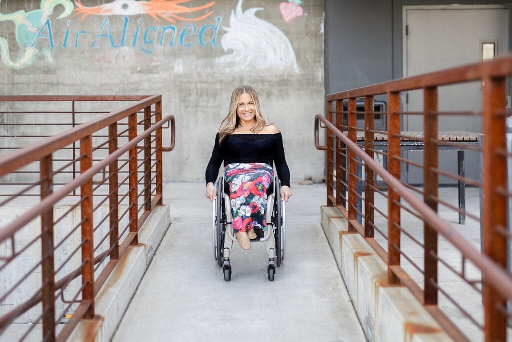 alycia wearing a black top and flower patterned skirt rolling down a ramp with painted red iron rails with a faded mural behind her