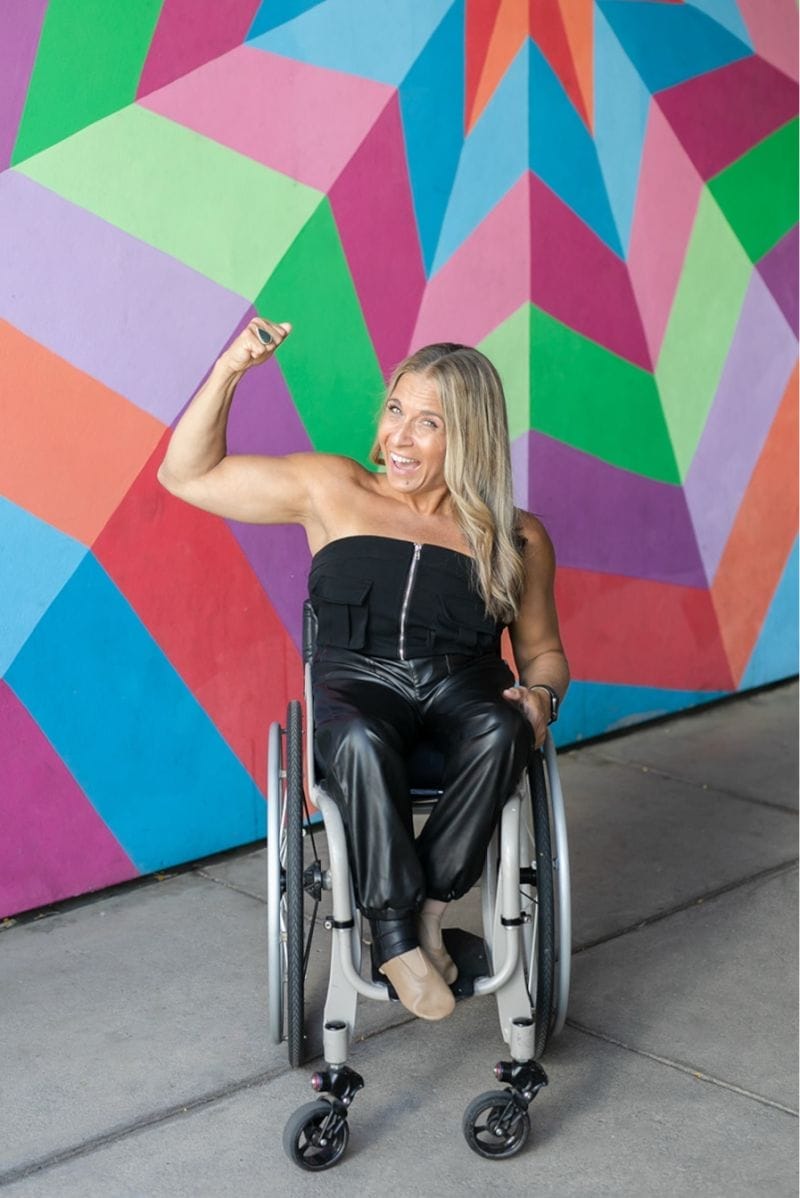 Alycia in front of a colorful wall wearing all back flexing her arm.