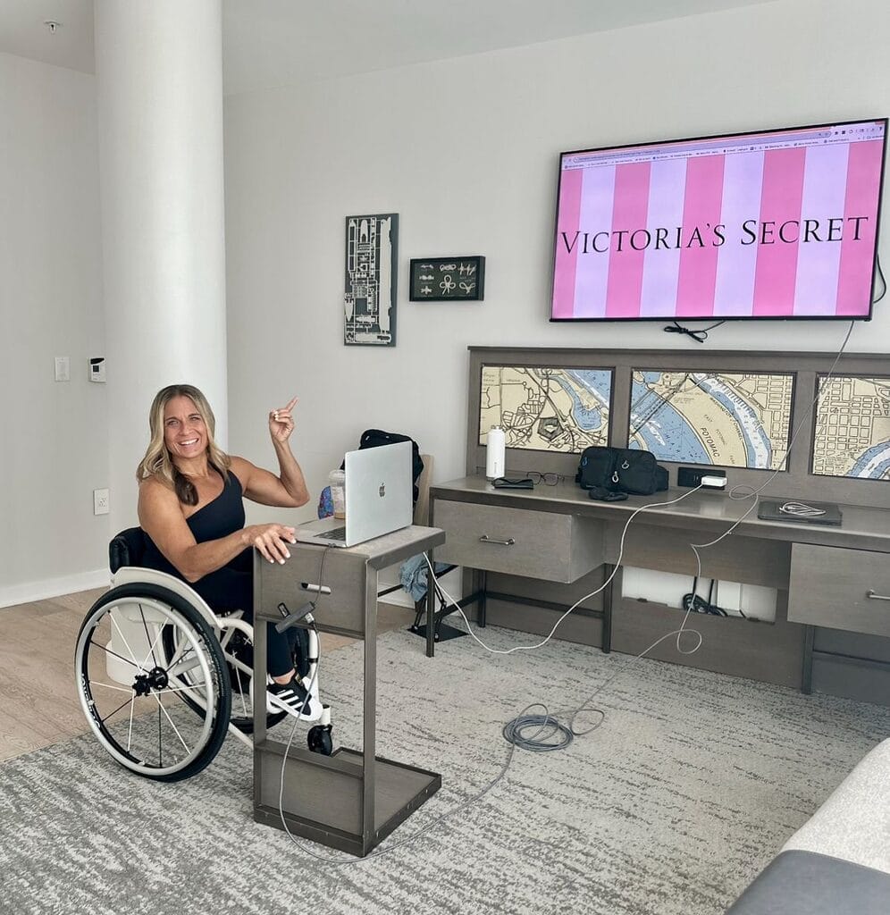 Alycia sitting in her wheelchair at a desk in front of two screens giving her keynote pointing at the top screen with Victoria's Secrete above. 